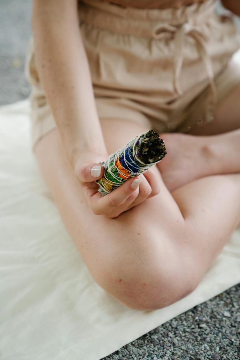 A woman meditatively holds a colorful sage smudge stick during a mindfulness practice outdoors.